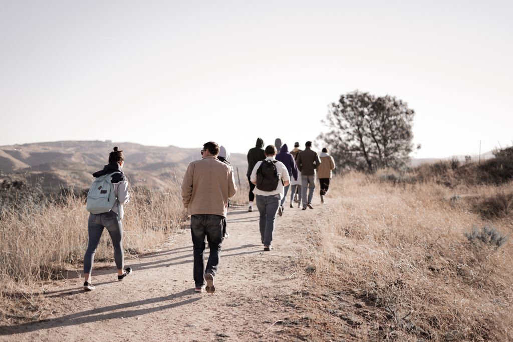 A group of adults hiking on a scenic, unpaved trail through dry grass and rolling hills on a sunny day.