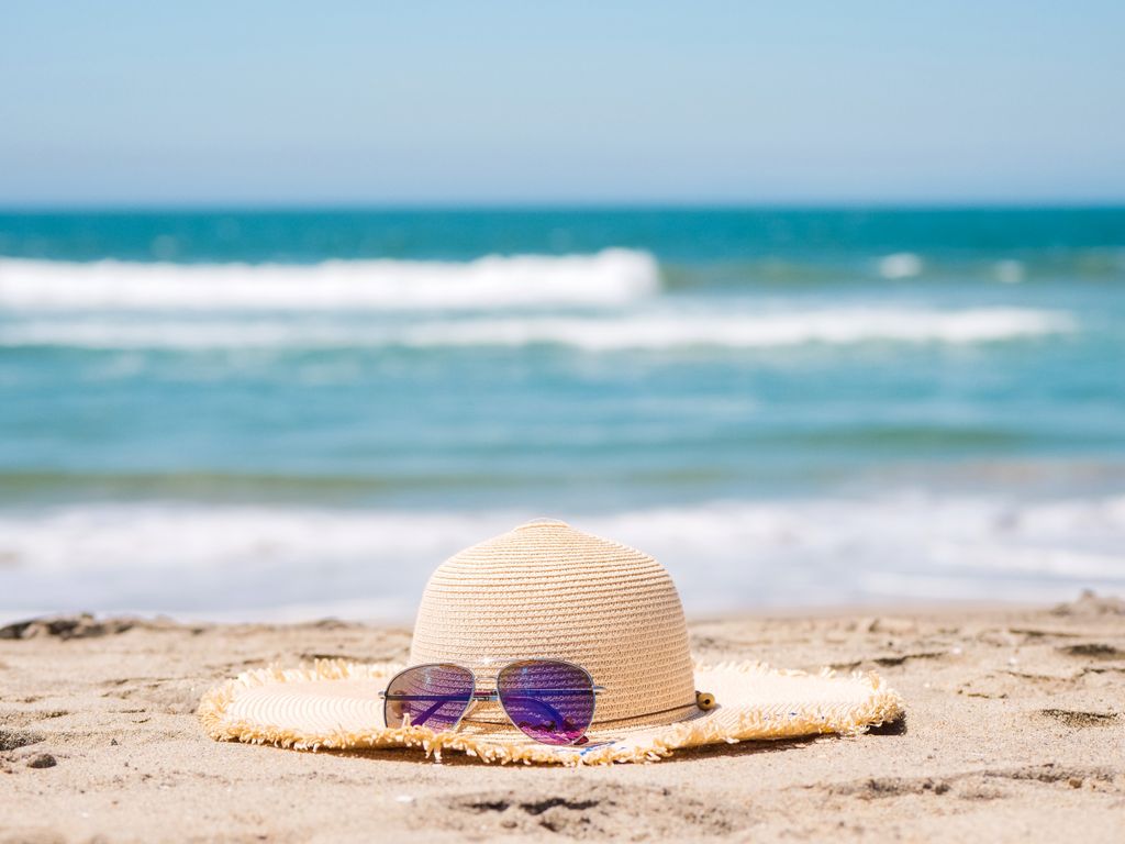 A straw hat and sunglasses resting on a sandy beach with blue waves in the background.